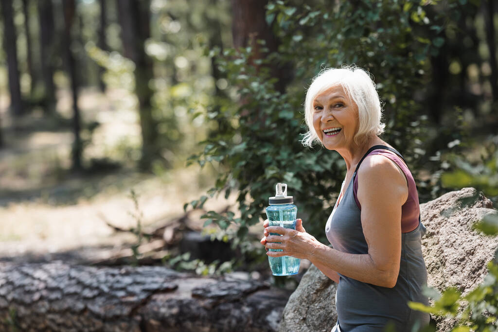 Веселая старшая спортсменка держит в лесу спортивную бутылку  - Фото, изображение