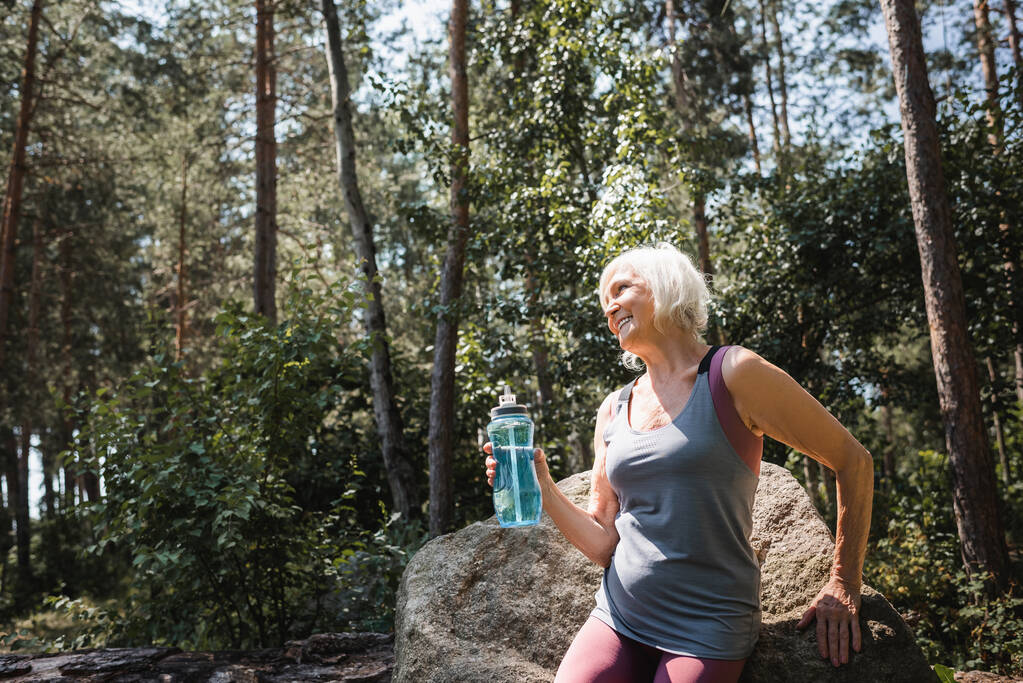 Fröhliche ältere Sportlerin hält Sportflasche in der Nähe von Stein im Wald  - Foto, Bild