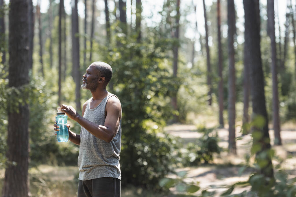 Πλευρική άποψη του ηλικιωμένου Αφροαμερικανού αθλητή κρατώντας αθλητικό μπουκάλι στο δάσος  - Φωτογραφία, εικόνα