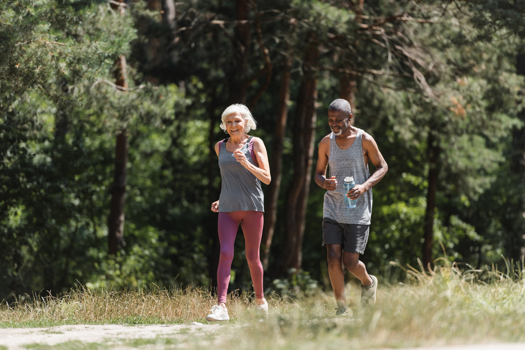 Femme sportive senior courir près de mari afro-américain avec bouteille de sport dans la forêt  - Photo, image