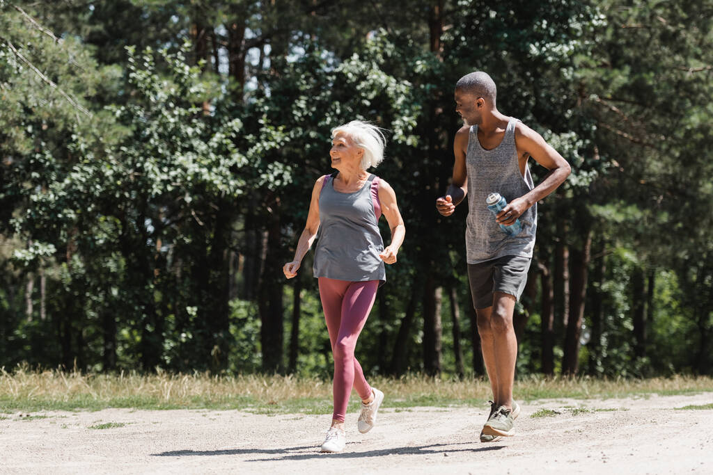 アフリカ系アメリカ人の夫の近くでスポーツボトルと一緒に森の中をジョギングする笑顔シニアスポーツ女性  - 写真・画像
