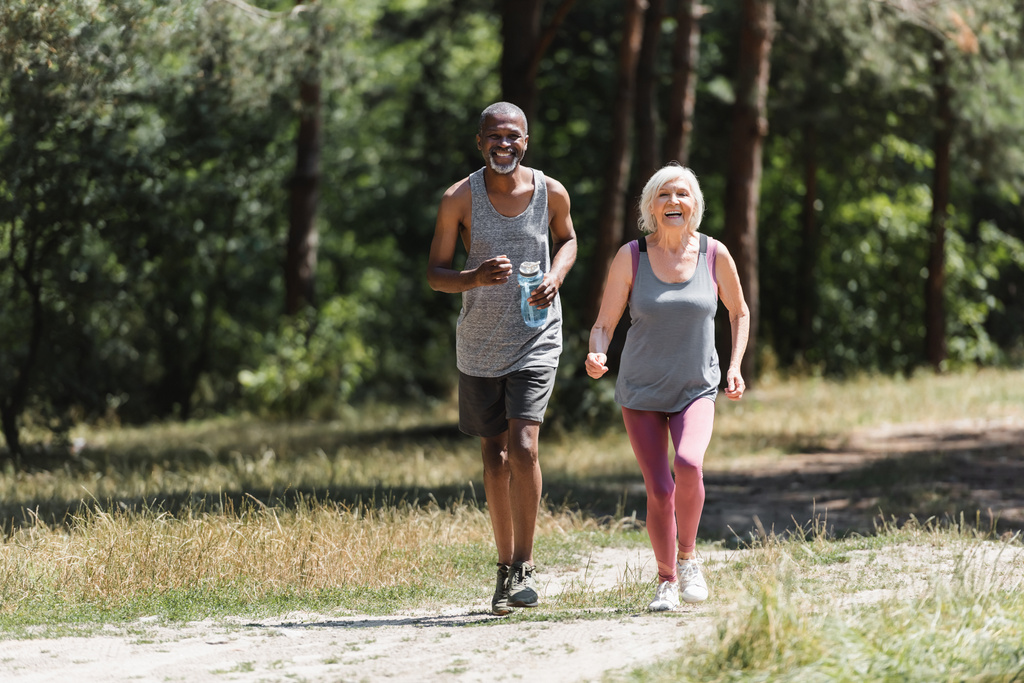 Spor şişeli Afrikalı Amerikalı sporcu ormanda mutlu bir eşin yanında koşuyor.  - Fotoğraf, Görsel