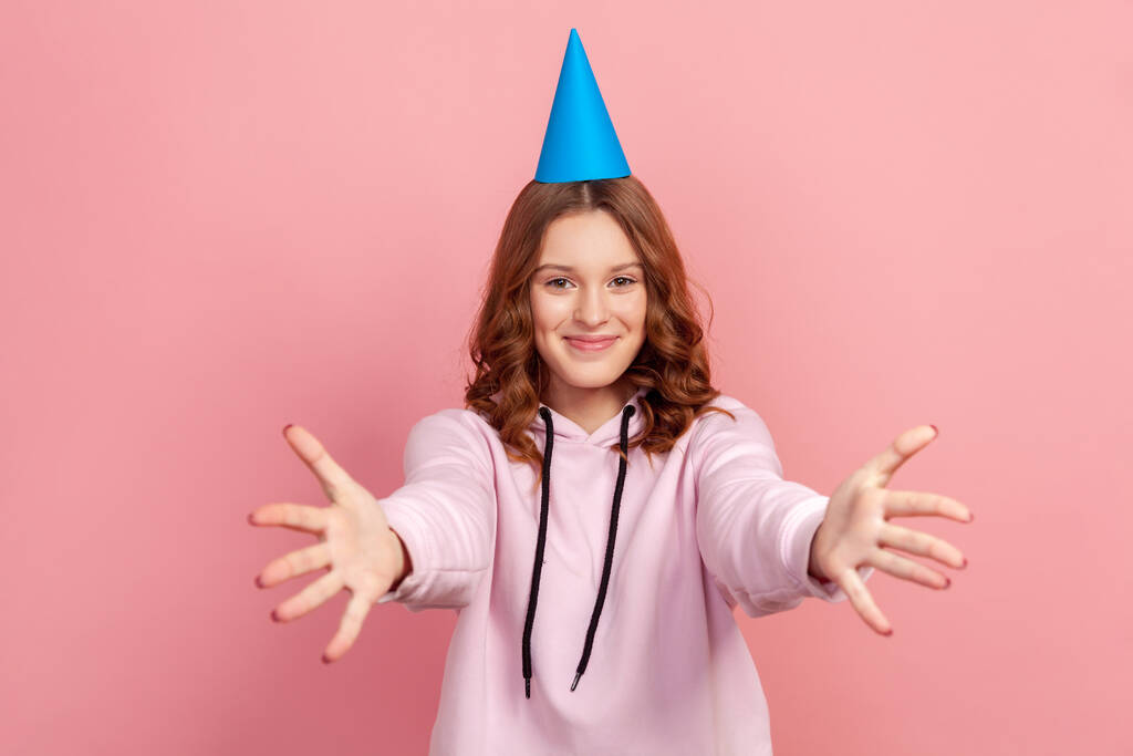 Portrét veselé kudrnaté puberťačky v kapuci s čepicí, která natahuje ruce, aby ji objala a usmála se a pozdravila. Vnitřní studio záběr izolované na růžovém pozadí - Fotografie, Obrázek
