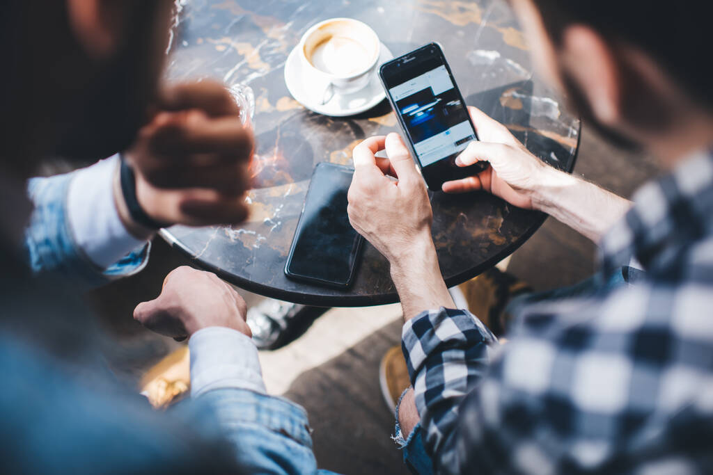 Урожай сверху вид современных парней в непринужденной стильной одежде, читающих сообщение на экране смартфона, отдыхающего в кафе - Фото, изображение