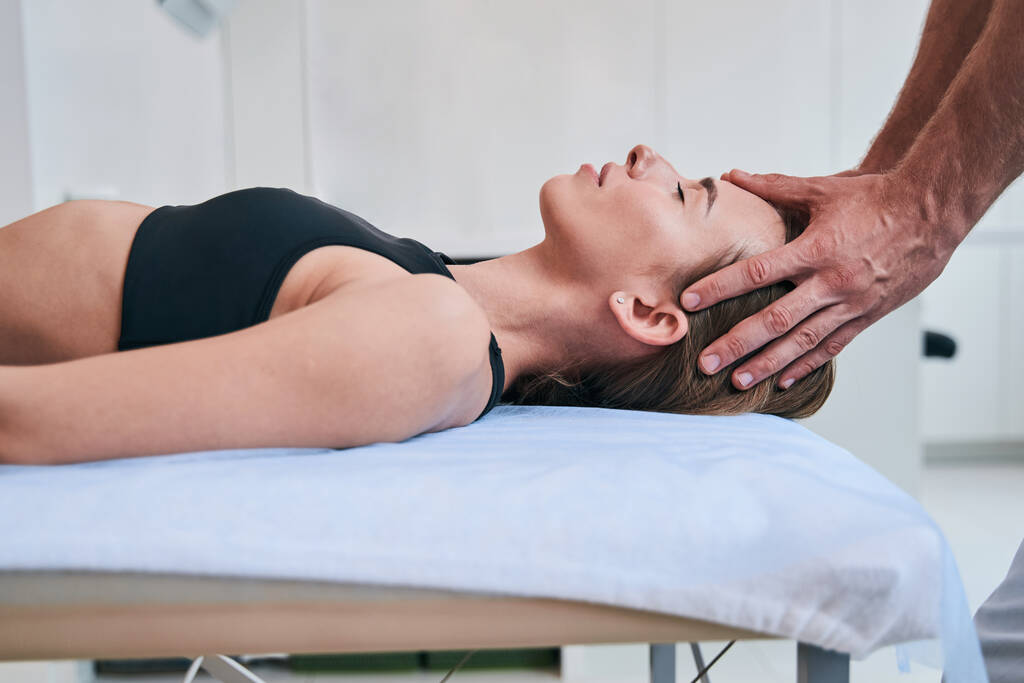Young elegant Caucasian woman in black sports bra enjoying facial massage in medical office - Photo, Image