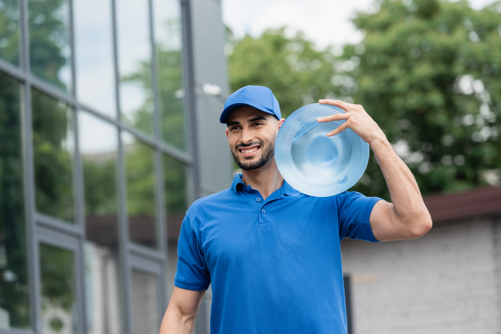 Wesoły arabski dostawca w czapce z butelką wody na zewnątrz  - Zdjęcie, obraz
