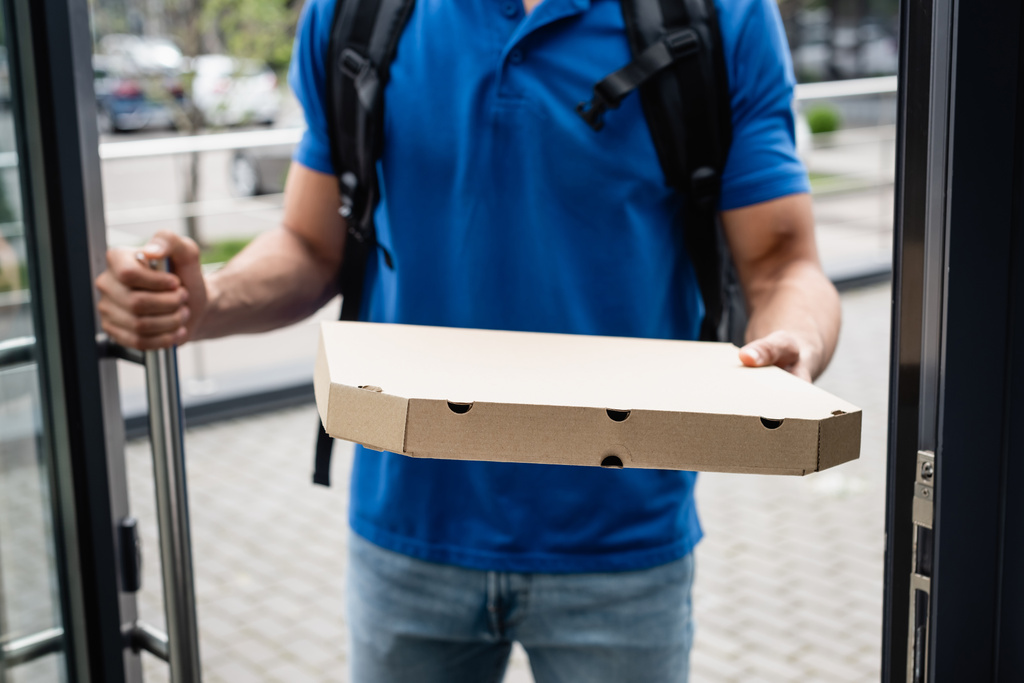 Vista recortada de la caja de pizza en la mano del mensajero borroso cerca de la puerta  - Foto, Imagen