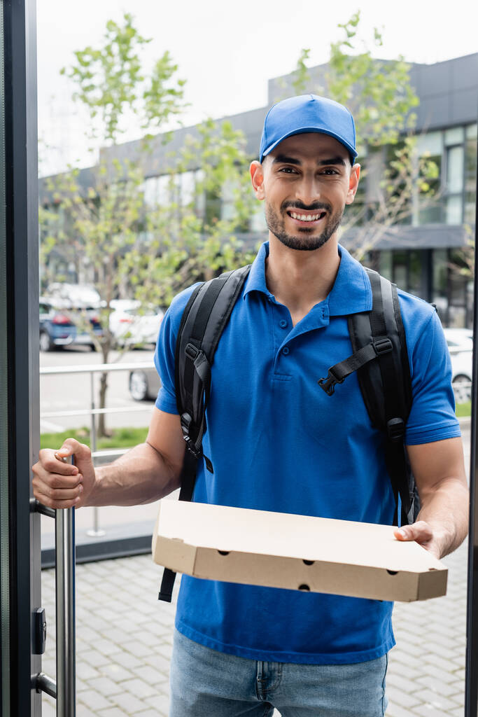 Fröhlicher muslimischer Lieferant hält Pizzaschachtel vor der Tür  - Foto, Bild