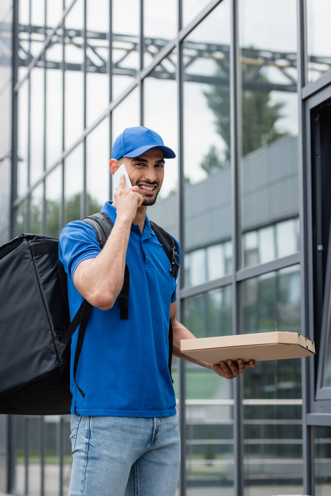 Usmívající se arabský doručovatel s batohem a pizza box mluví na smartphone venku  - Fotografie, Obrázek