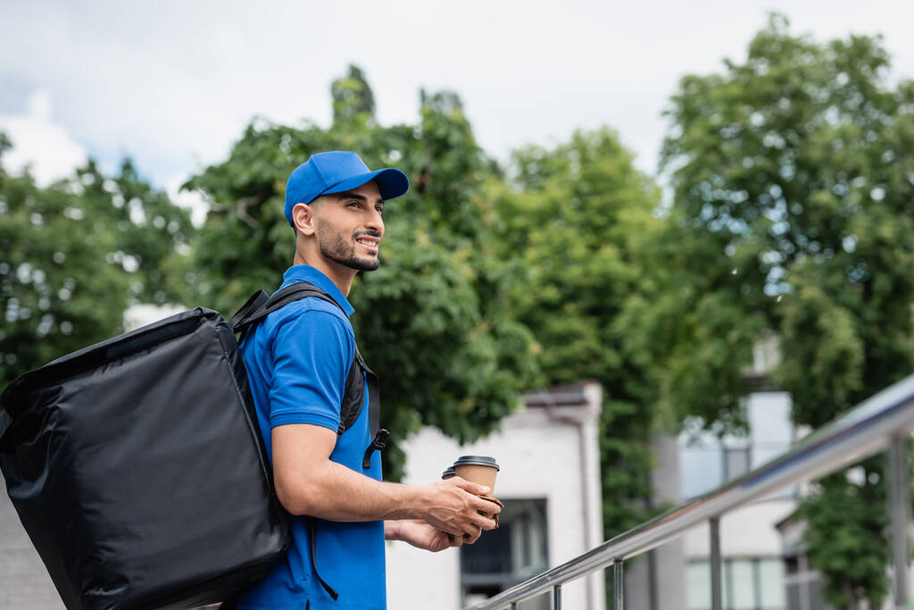 Smiling arabian courier with thermo backpack holding coffee cups outdoors  - Photo, Image