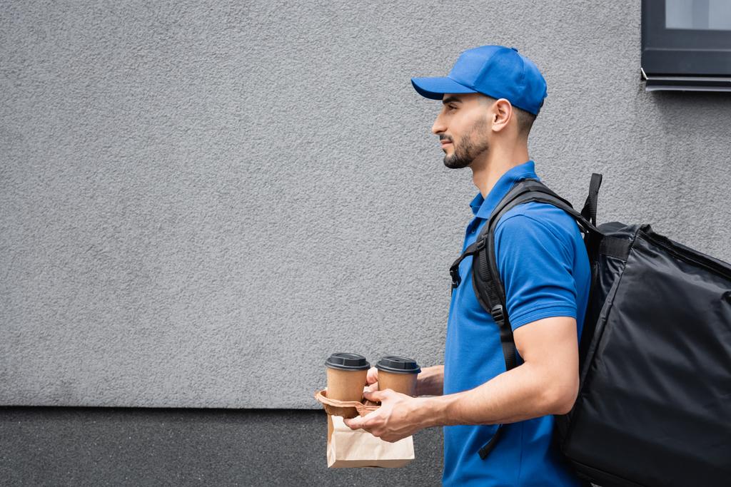 Side view of arabian deliveryman holding paper cups and bag near building  - Photo, Image
