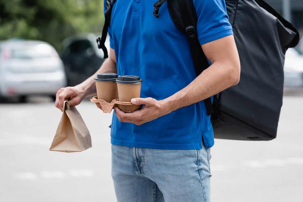 Обрезанный вид курьера с терморюкзаком держа бумажные стаканчики и сумки на открытом воздухе  - Фото, изображение