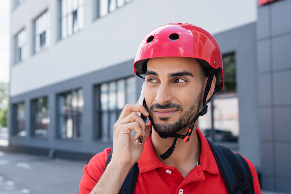 Мусульманский курьер в шлеме с рюкзаком разговаривает по смартфону на улице  - Фото, изображение