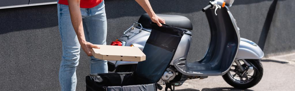 Vista recortada del repartidor sosteniendo la caja de pizza cerca de la mochila termo abierta y el scooter al aire libre, pancarta  - Foto, imagen