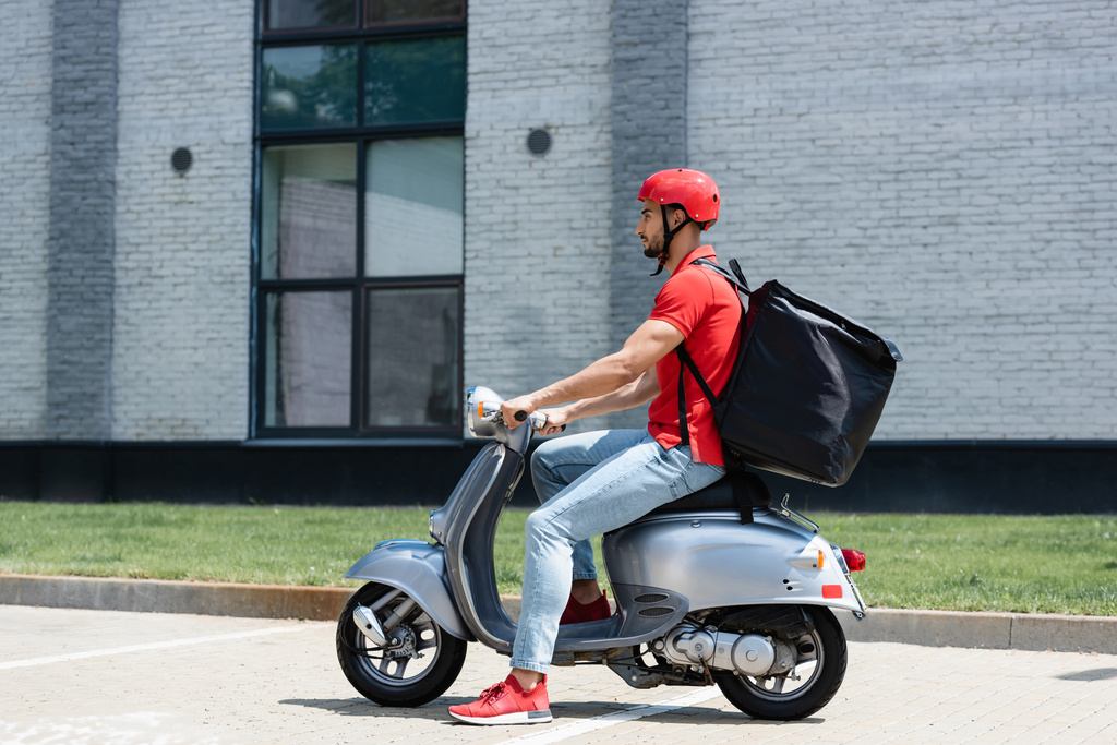 Vue latérale du courrier musulman en uniforme rouge et sac à dos thermique scooter d'équitation à l'extérieur  - Photo, image