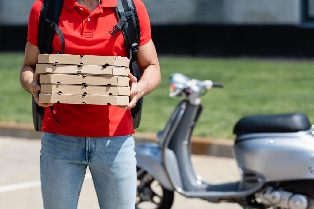 Vue recadrée du livreur avec sac à dos thermique tenant des boîtes à pizza près du scooter flou à l'extérieur  - Photo, image