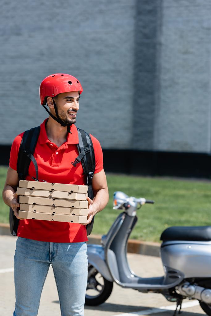 Alegre árabe entregador segurando caixas de pizza perto de scooter borrado ao ar livre  - Foto, Imagem