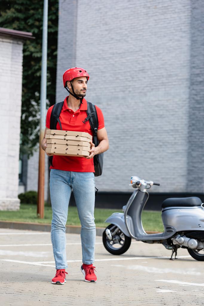 Correio muçulmano em capacete segurando caixas de pizza perto de scooter borrado ao ar livre  - Foto, Imagem