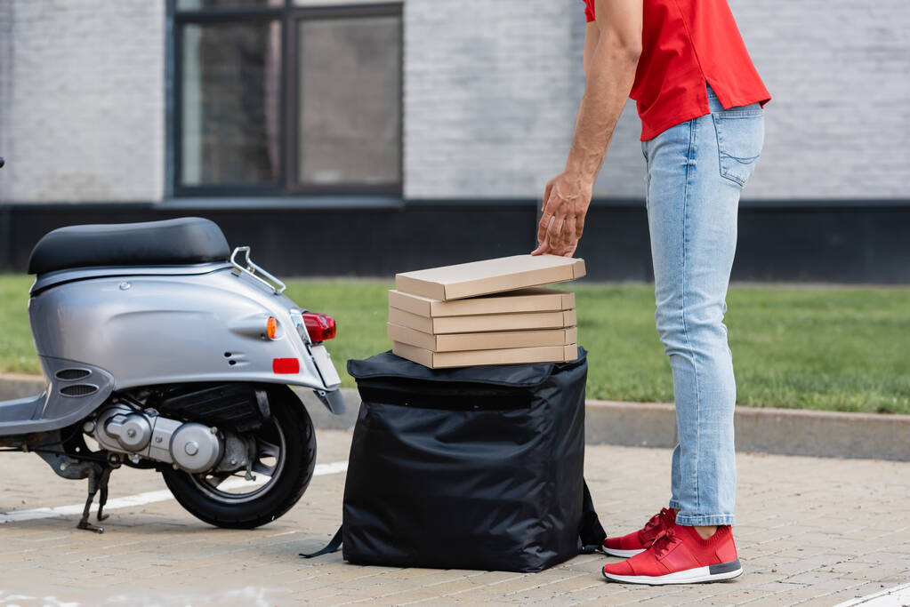 Vue recadrée du courrier mettant des boîtes à pizza sur le sac à dos thermique près du scooter à l'extérieur  - Photo, image