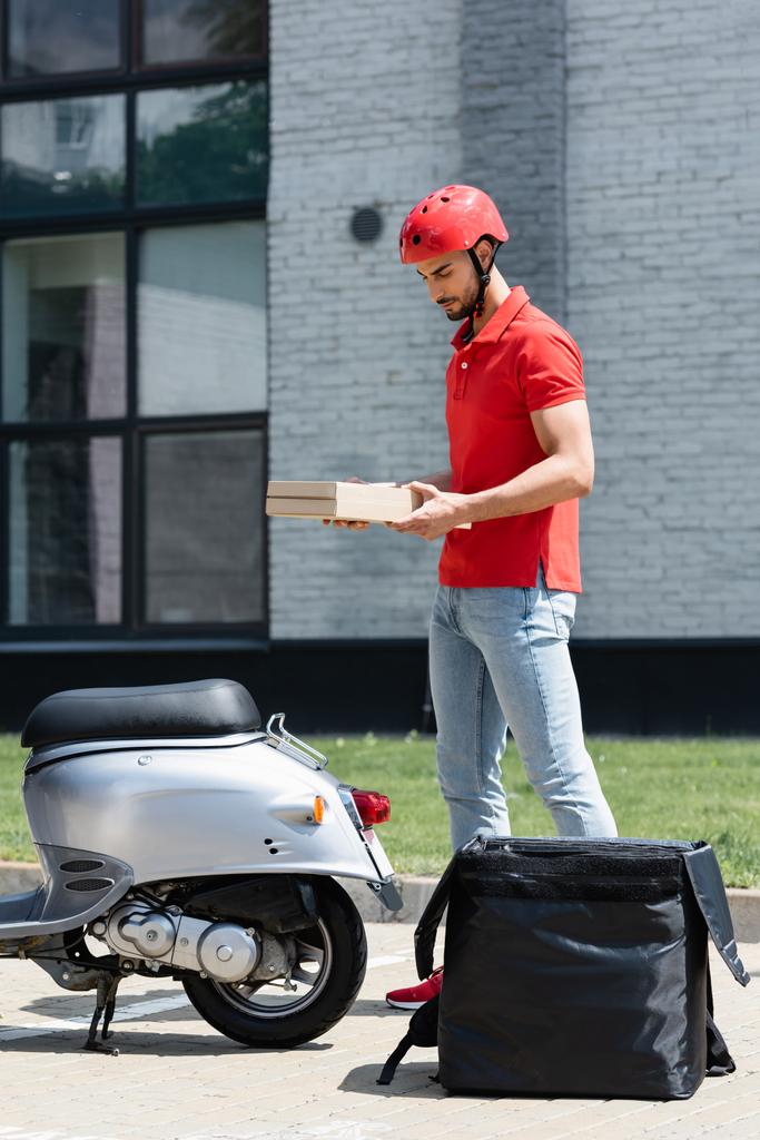 Jeune messager musulman dans un casque tenant des boîtes à pizza près de scooter et sac à dos thermo à l'extérieur  - Photo, image