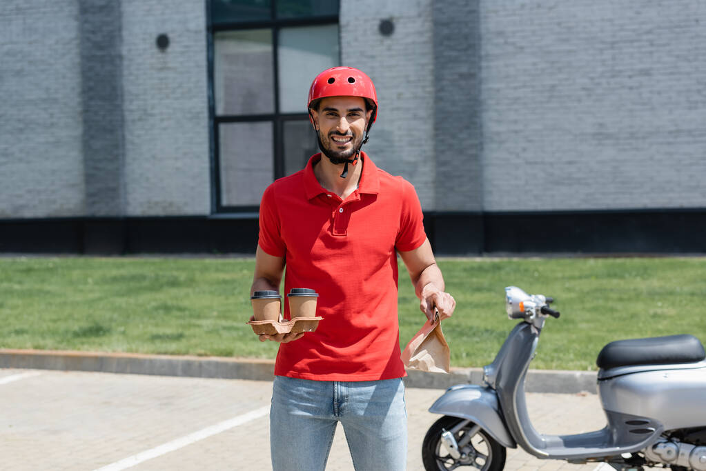 Smiling muslim deliveryman in helmet holding coffee to go and paper bag near blurred scooter outdoors  - Photo, Image