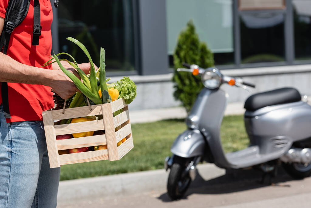 Cropped view of deliveryman holding wooden box with fresh vegetables near blurred scooter outdoors  - Photo, Image