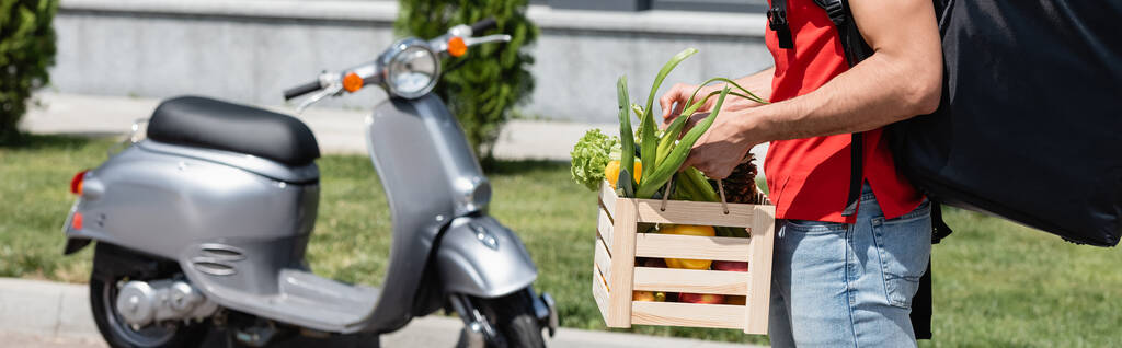 Καλλιεργημένη άποψη του deliveryman με λαχανικά και σακίδιο στέκεται κοντά θολή σκούτερ σε εξωτερικούς χώρους, πανό  - Φωτογραφία, εικόνα