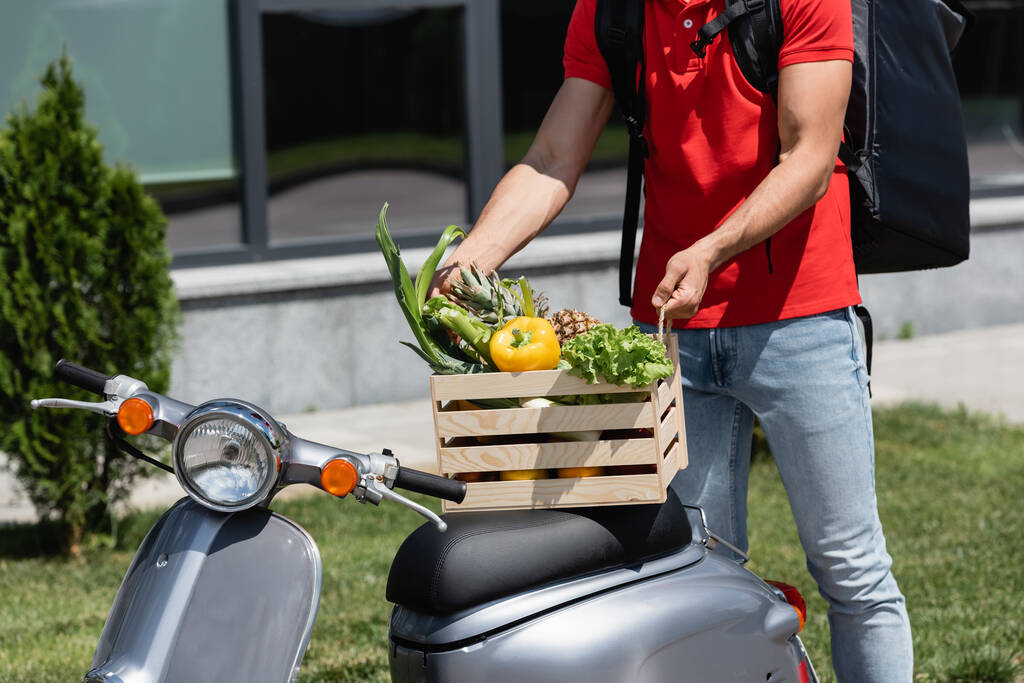 Teslimatçının elinde sebzeli ahşap kutu ve dışarıda scooterlı ananas tuttuğu görüntüler.  - Fotoğraf, Görsel