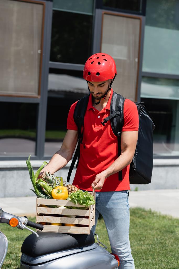 Junger arabischer Kurier im Schutzhelm hält Holzkiste mit Gemüse auf Roller  - Foto, Bild