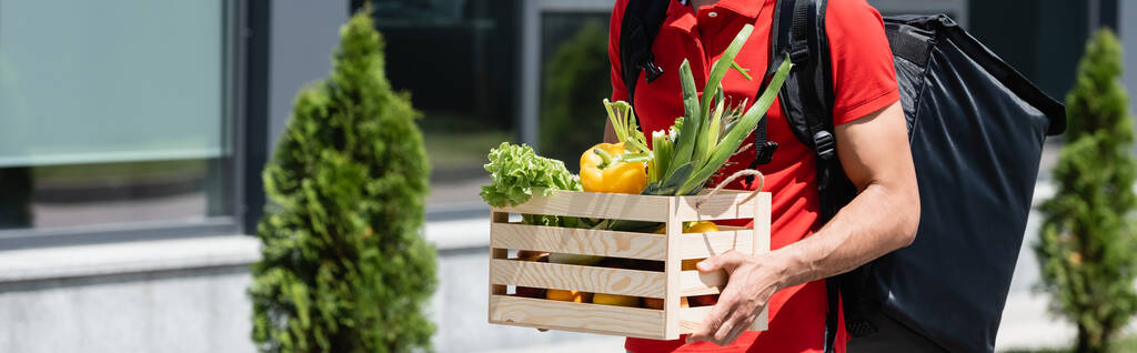 Vue recadrée du livreur tenant une boîte en bois avec des légumes à l'extérieur, bannière  - Photo, image
