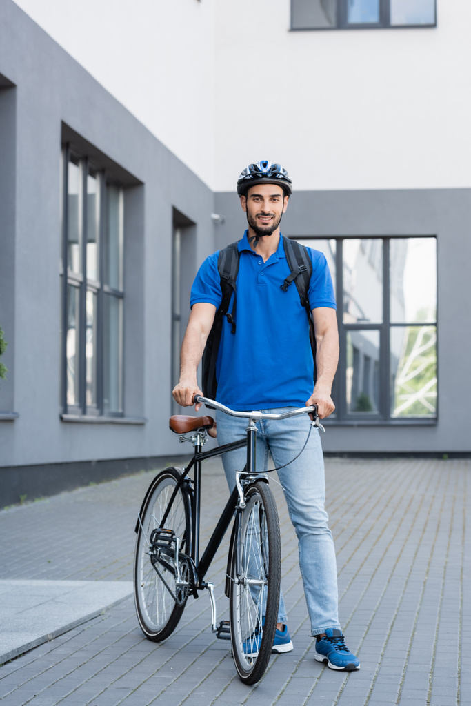 Courrier arabe souriant avec sac à dos debout près du vélo à l'extérieur  - Photo, image