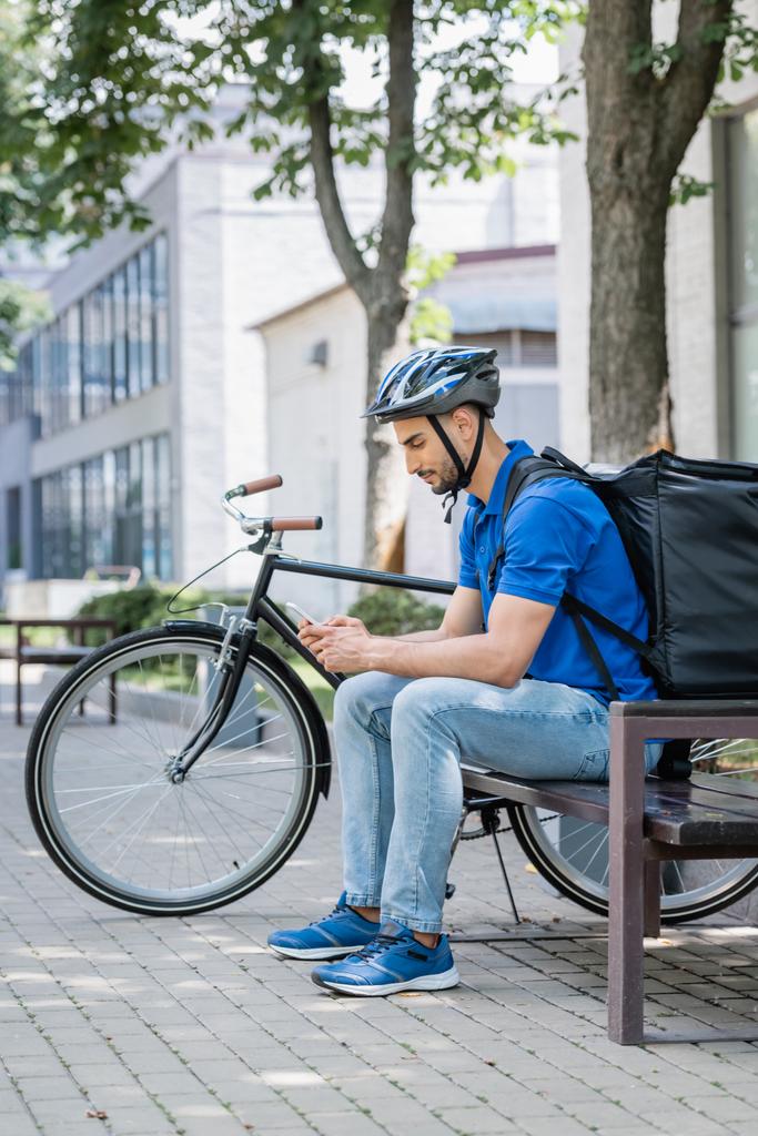 Seitenansicht muslimischer Kurier in blauem T-Shirt und Thermo-Rucksack mit Handy auf Bank neben Fahrrad  - Foto, Bild