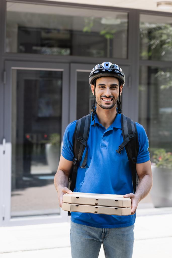 Lächelnder muslimischer Kurier mit Schutzhelm und Rucksack hält Pizzakartons in der Hand und blickt in die Kamera im Freien  - Foto, Bild