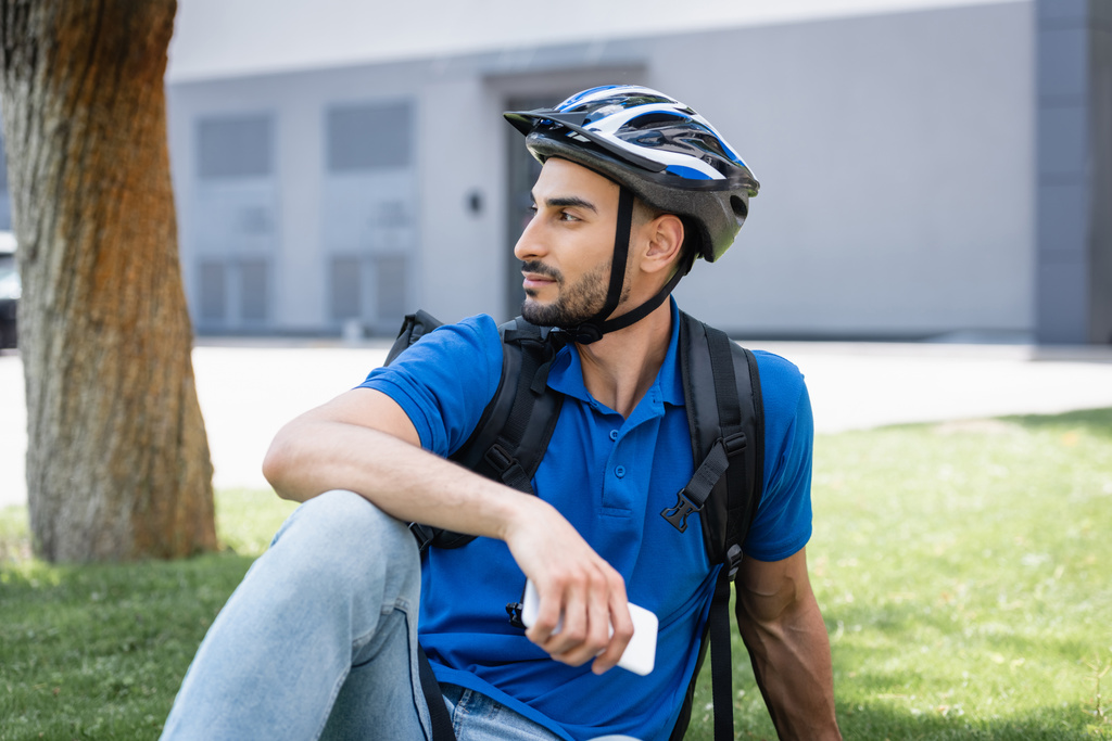 Vista lateral del repartidor musulmán en casco protector que sostiene el teléfono inteligente en el césped  - Foto, imagen