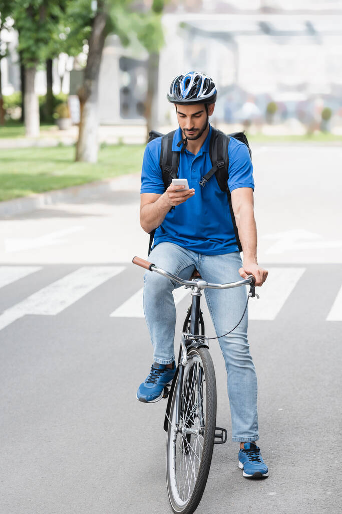 Repartidor árabe con mochila usando teléfono inteligente mientras monta bicicleta al aire libre  - Foto, imagen