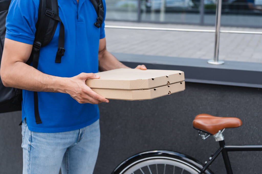 Καλλιεργημένη άποψη του courier κρατώντας κουτιά πίτσα κοντά θολή ποδήλατο σε εξωτερικούς χώρους  - Φωτογραφία, εικόνα