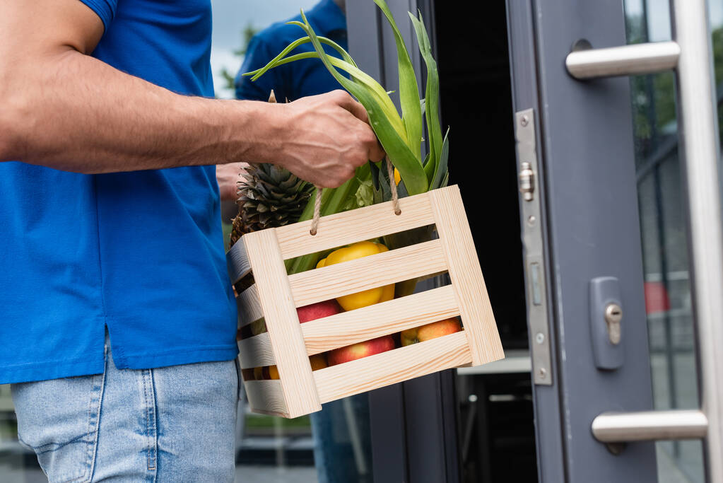 Vista cortada de entregador segurando caixa com alimentos frescos perto da porta embaçada  - Foto, Imagem