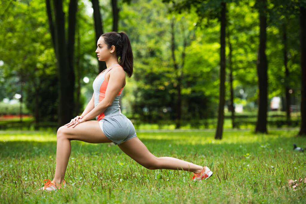 side view of pretty sportswoman doing lunge exercise in park  - Photo, Image