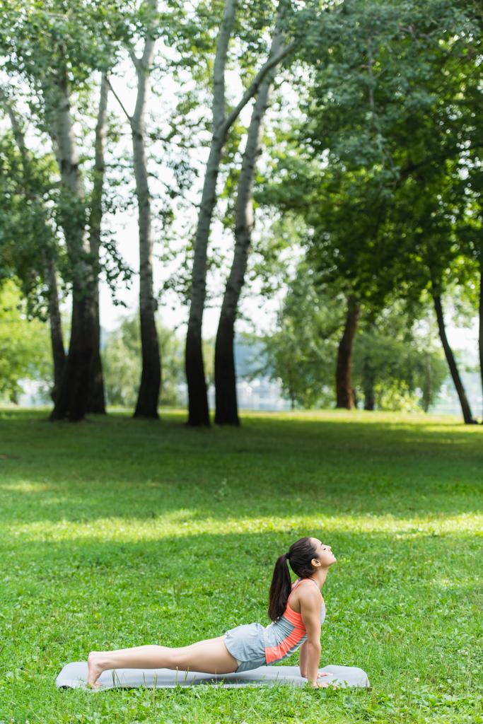 πλαϊνή άποψη του ταιριάζει γυναίκα με κλειστά μάτια που εκτείνεται σε στρώμα γιόγκα στο πάρκο - Φωτογραφία, εικόνα