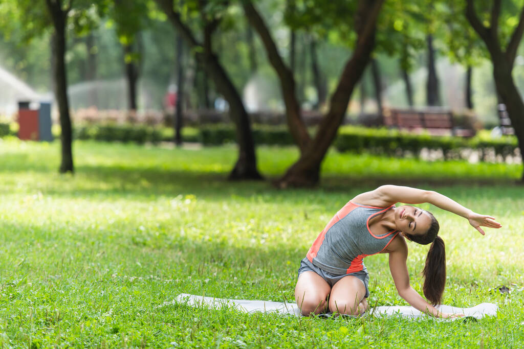 rugalmas nő sportruházatban nyújtózkodik jóga szőnyegen a parkban - Fotó, kép