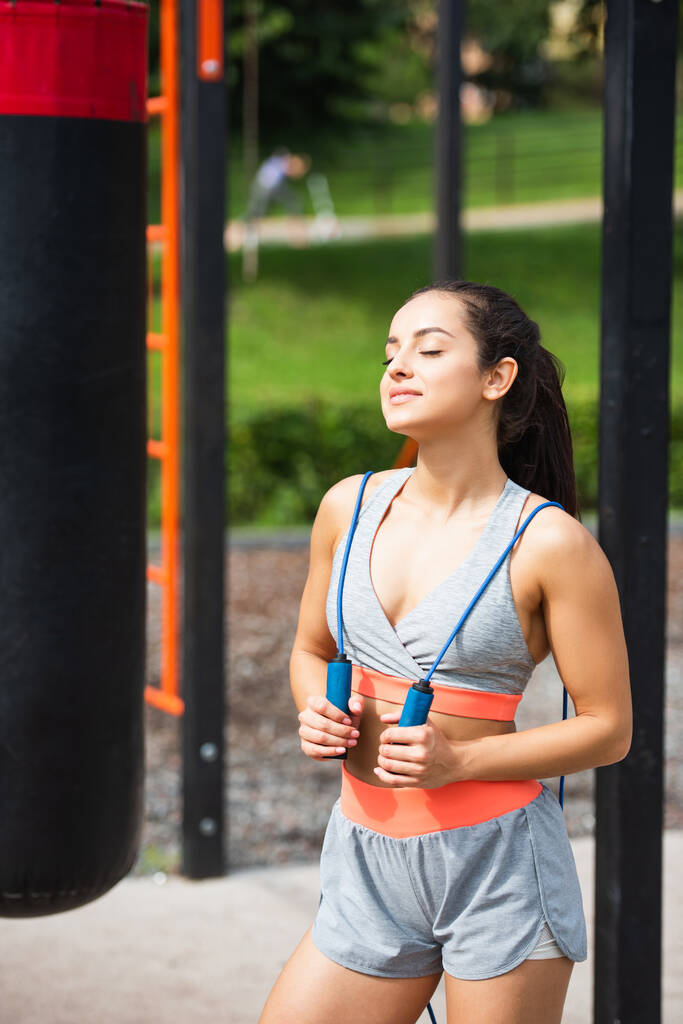 pleased sportswoman with closed eyes holding jumping rope outside  - Photo, Image