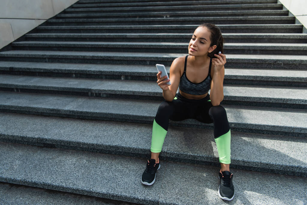 pleased sportswoman in wireless earphones using smartphone while listening music and sitting on stairs outside  - Photo, Image