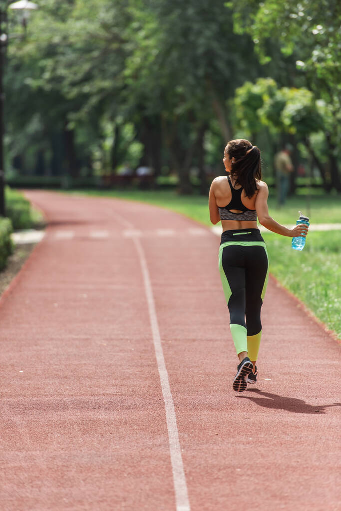 zpět pohled sportovkyně v obilí top a kamaše držící sportovní vodu při běhu v parku - Fotografie, Obrázek