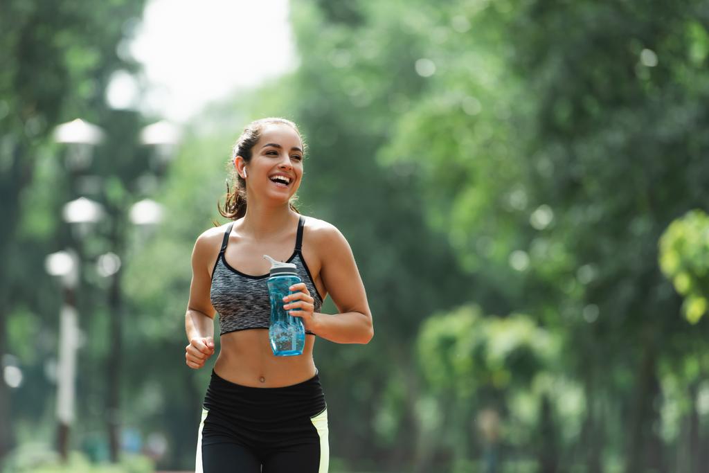 wesoły sportowiec w słuchawkach bezprzewodowych trzymając butelkę sportową podczas biegania w zielonym parku - Zdjęcie, obraz