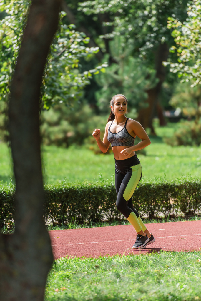pełna długość młodej sportowca w słuchawkach bezprzewodowych biegających po ścieżce w zielonym parku - Zdjęcie, obraz