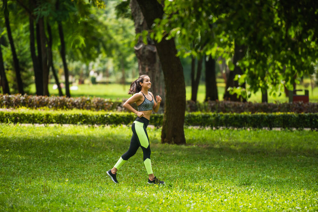pełna długość szczęśliwy sportowiec w górę upraw i legginsy jogging na trawie w parku  - Zdjęcie, obraz