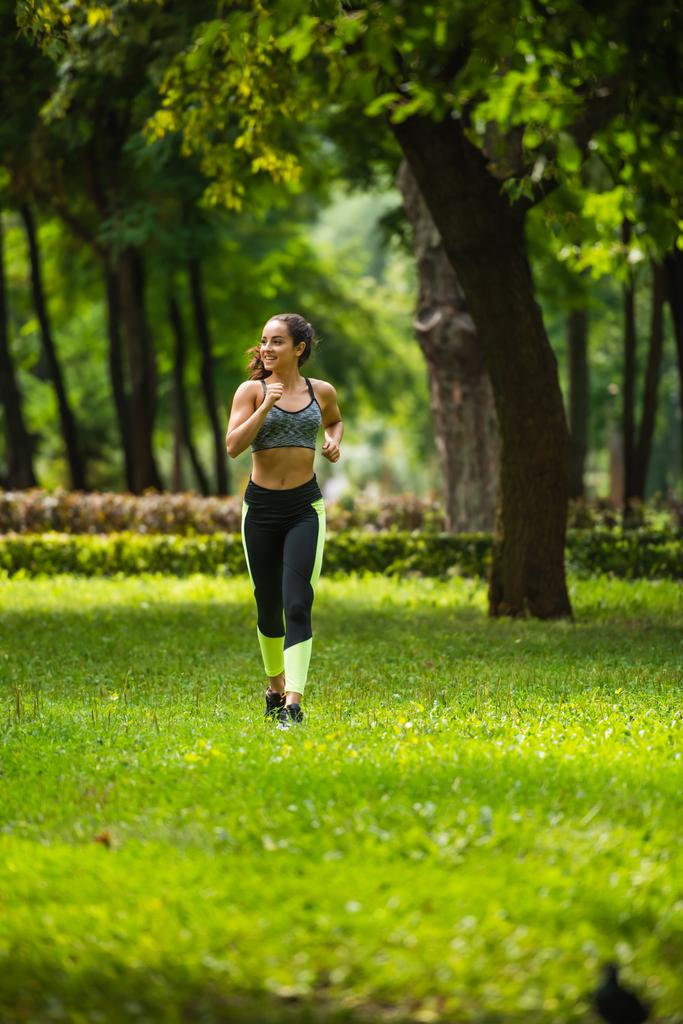 volledige lengte van glimlachende sportvrouw in gewas top en leggings joggen op gras in park  - Foto, afbeelding
