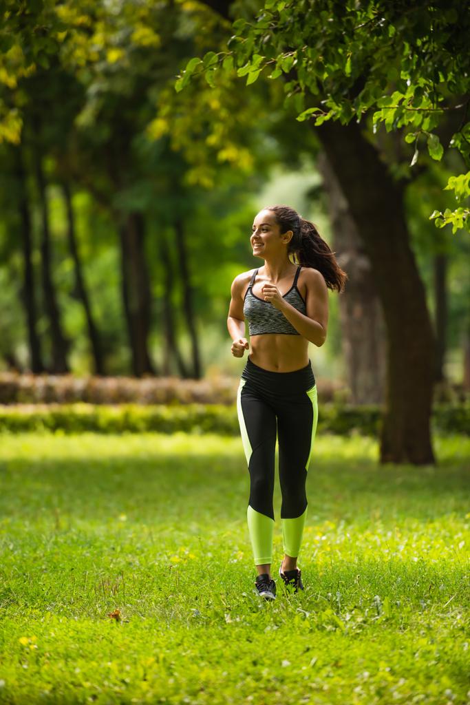 volledige lengte van positieve sportvrouw in gewas top en leggings joggen op gras in park  - Foto, afbeelding