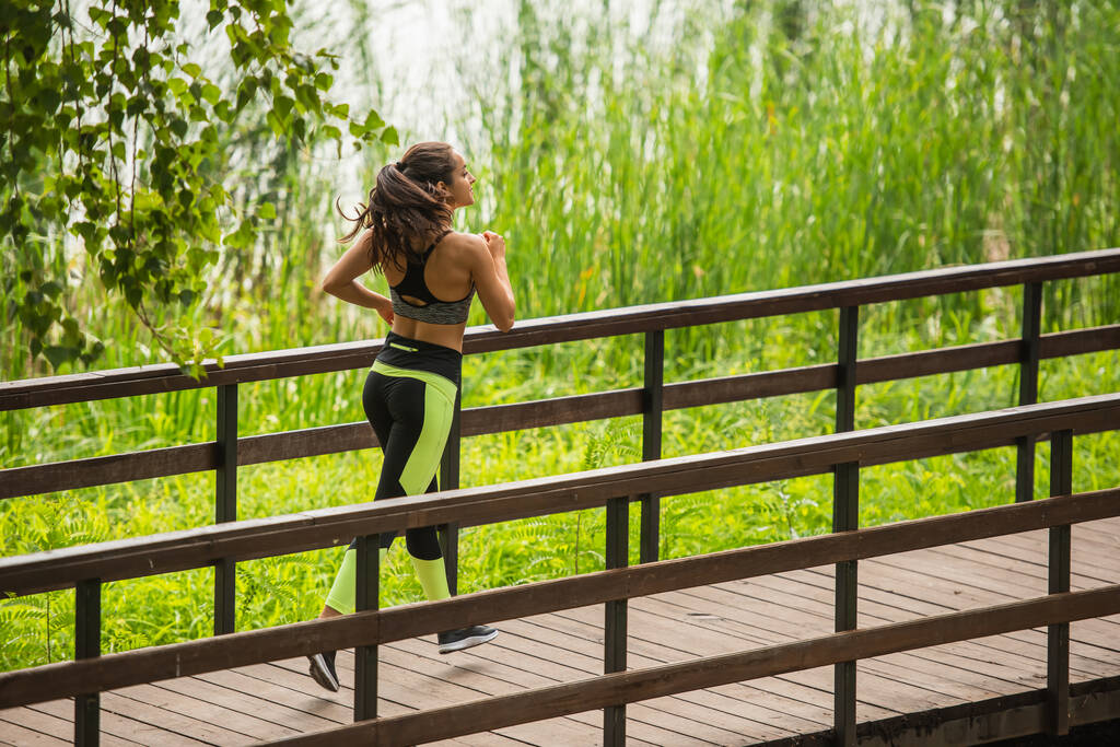volle Länge der glücklichen Sportlerin in Crop Top und Leggings beim Joggen auf Brücke im Park  - Foto, Bild