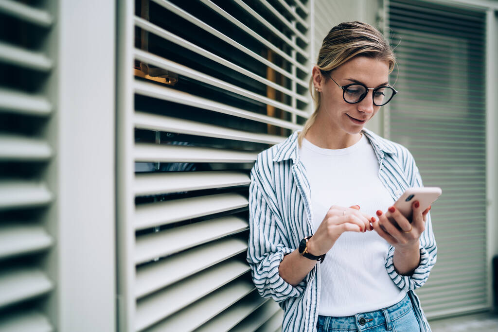 Lächelnde junge schöne Dame in lässiger Kleidung und trendiger Brille sucht mit dem Smartphone nach Informationen, während sie sich an die Jalousie lehnt - Foto, Bild